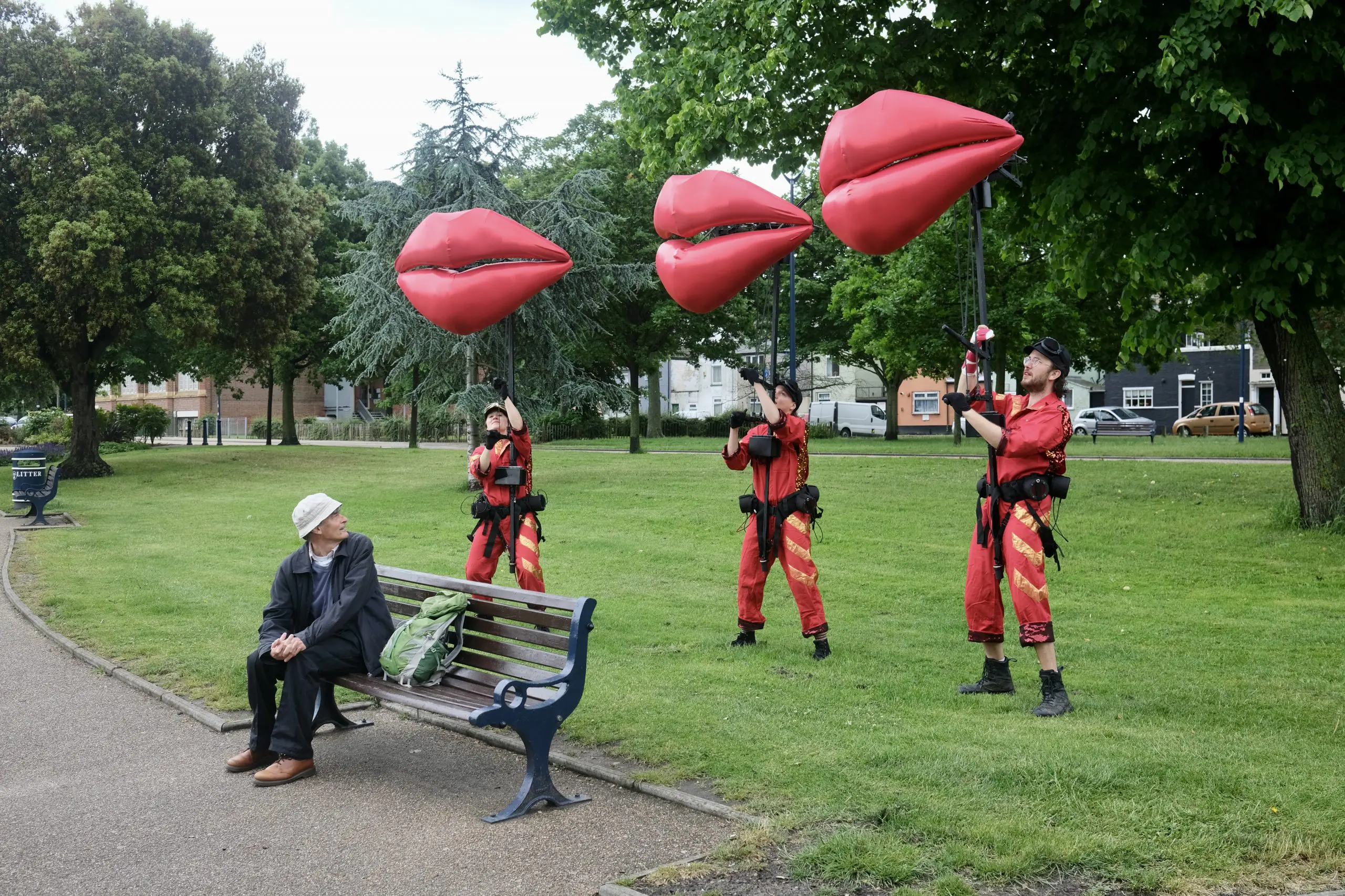 Three puppeteers with the giant red lips models, standing on grass behind a person on a bench, who has turned to watch them.