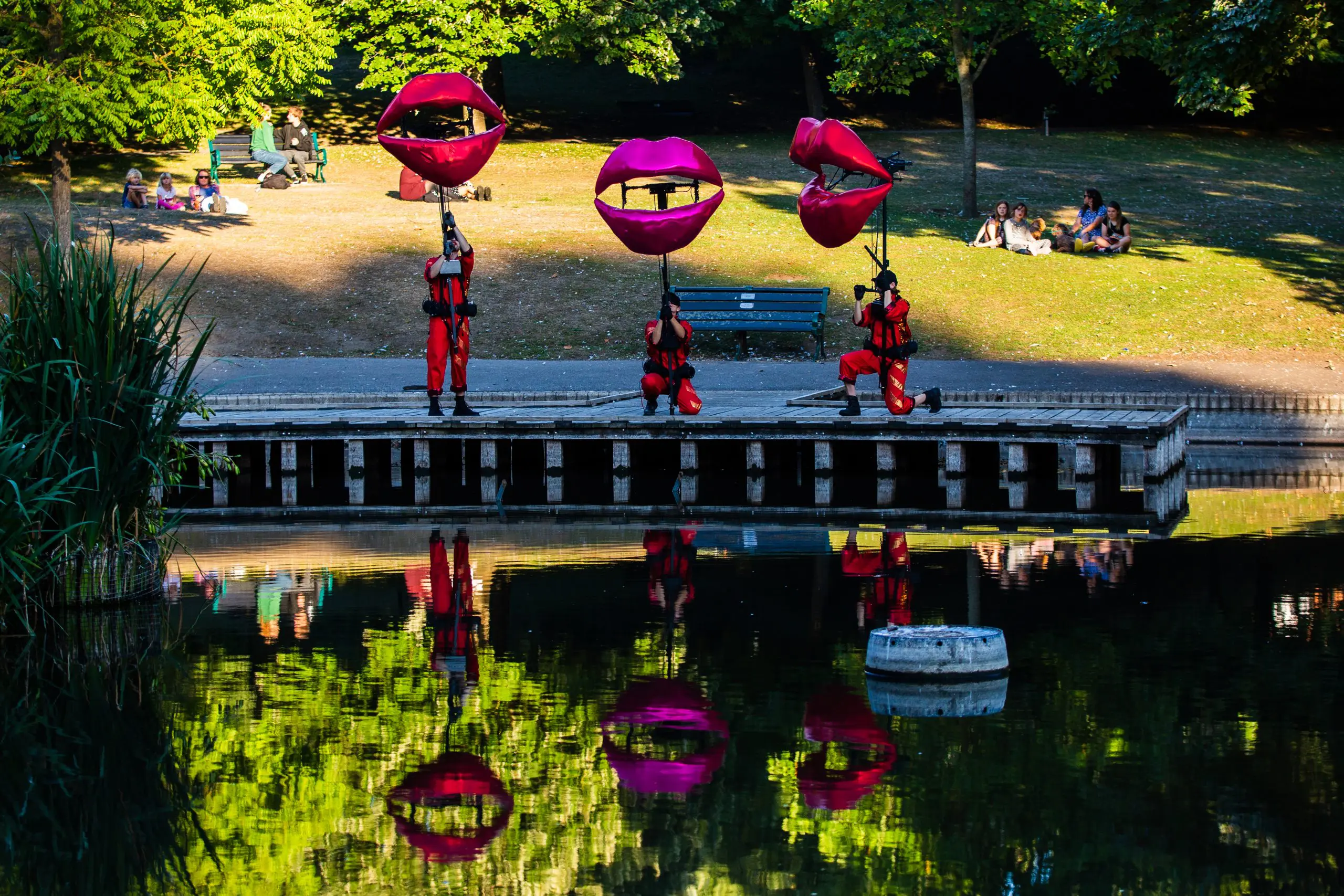 Three large puppets shaped like pairs of red lips. The puppeteers stand on a promenade and are reflected in the water in front of them.