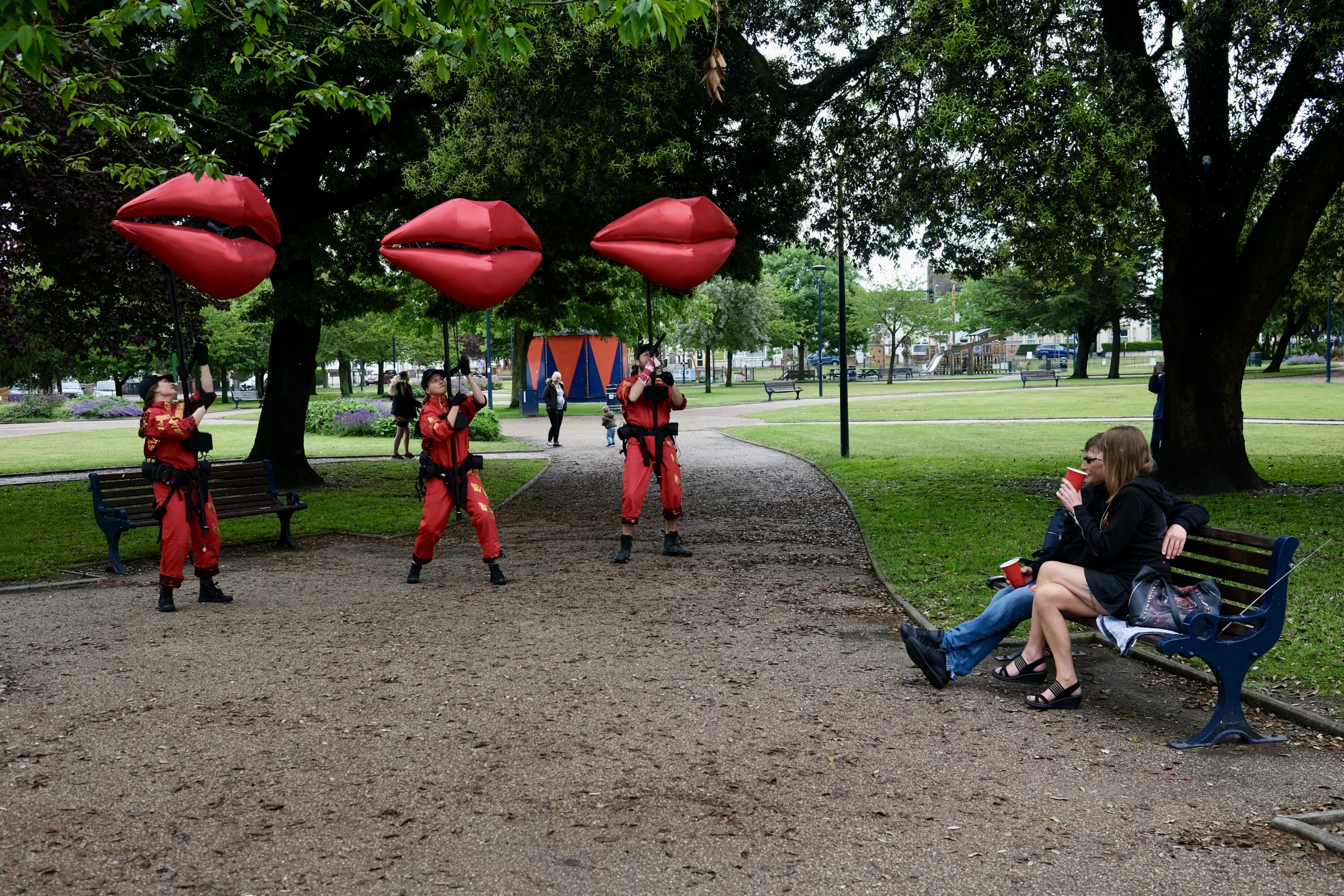 Three puppets manned by three puppeteers, of giant red pairs of lips. Two people sit on a bench opposite them.