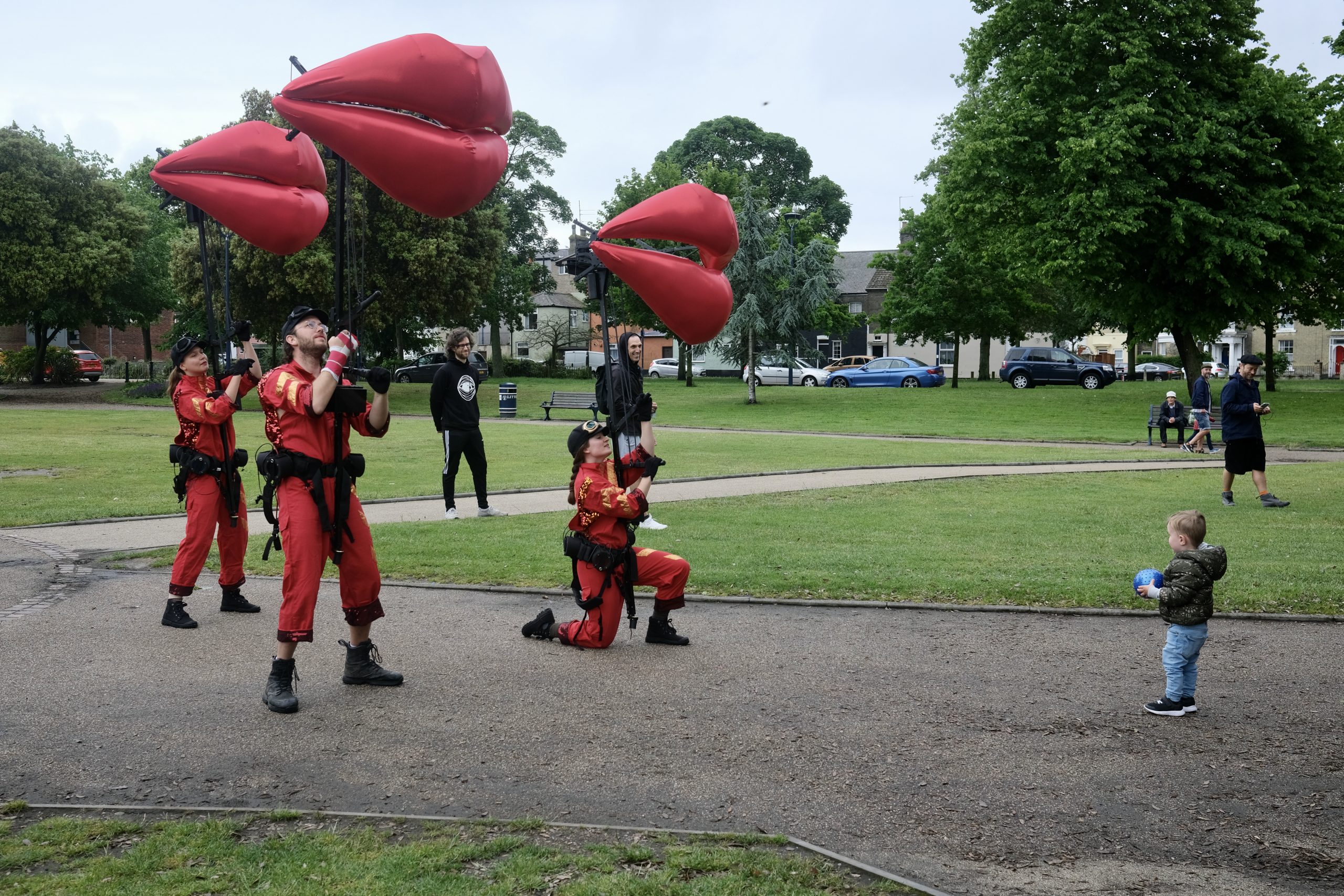 Three puppeteers with models of giant red pairs of lips, one kneeling, in front of a toddler in a park.