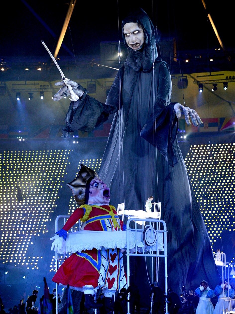 A giant puppet, 40-feet tall, resembling 'Voldemort' from the 'Harry Potter' series, holding a giant wand. It's 'head' is pointed towards a 20-foot tall puppet of a queen, frowning with her mouth wide open. She's in a hospital bed next to a real-life-sized hospital bed, suspended in midair with a child sitting up in it. Below all this on the ground is a crowd of performers.