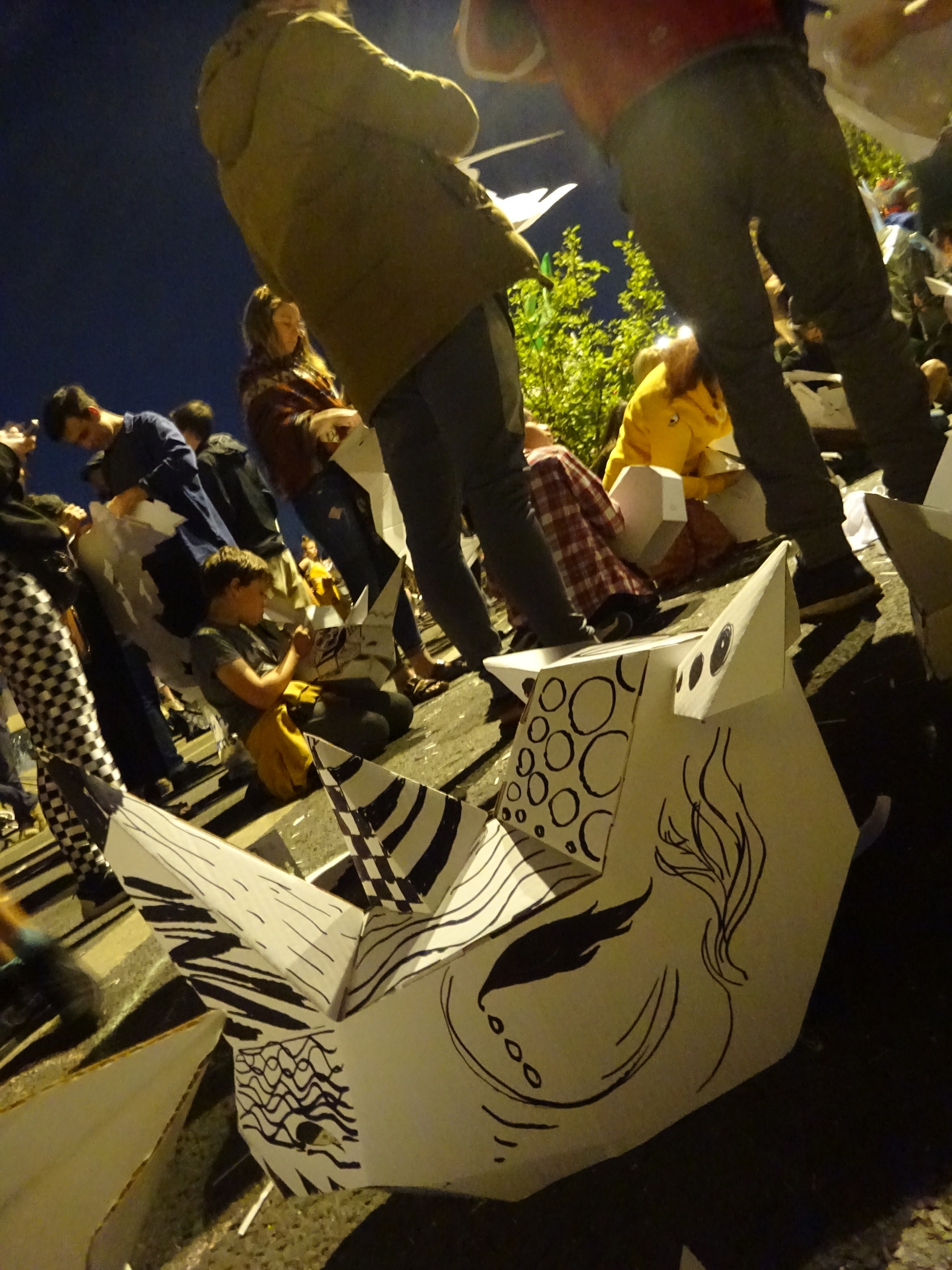 A white card rhino mask, decorated with patterns in black pen. It is on the ground, with people behind it decorating their own masks.