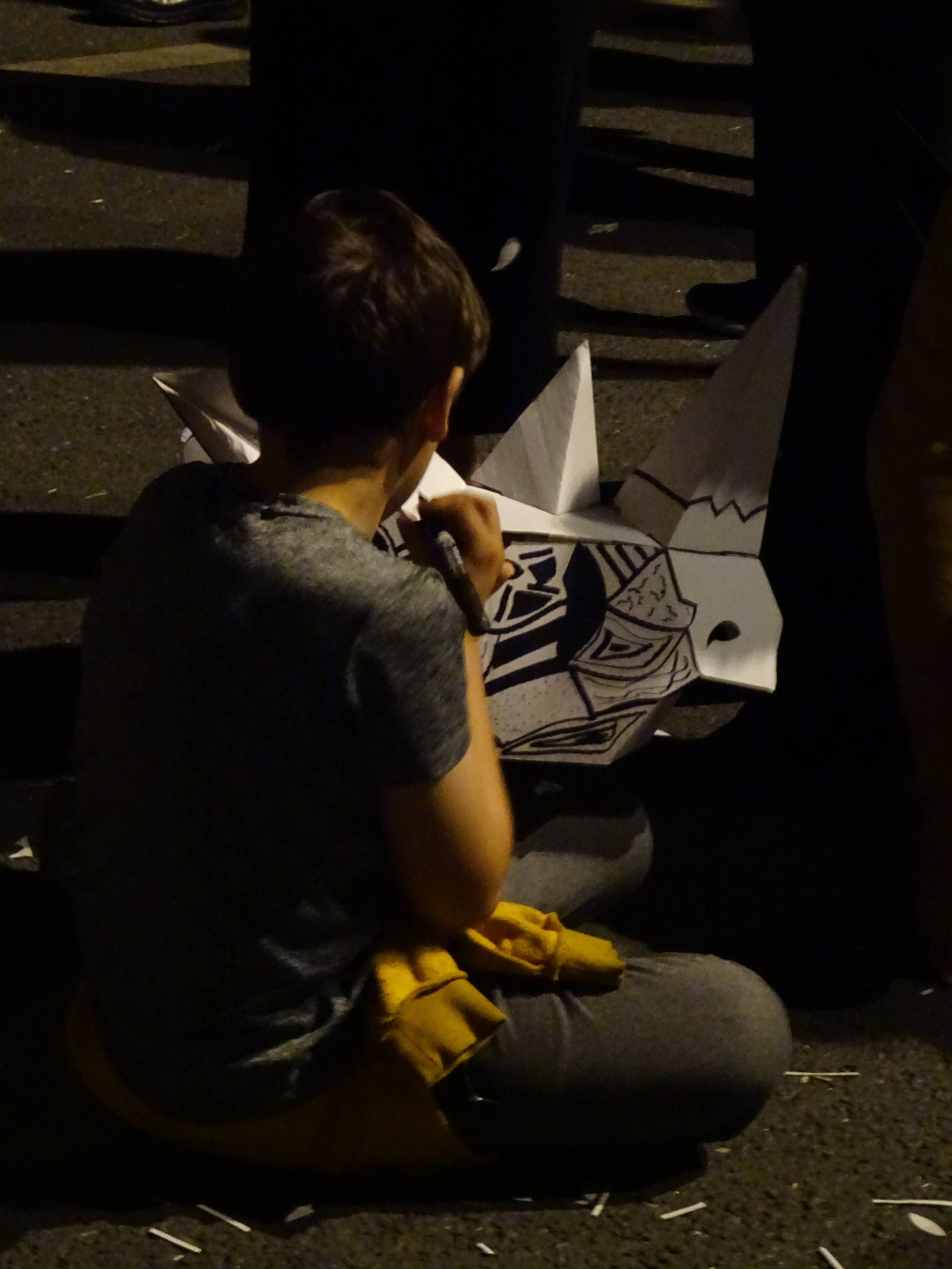 A person sits on the ground, decorating a rhino mask with black pen. It's night.