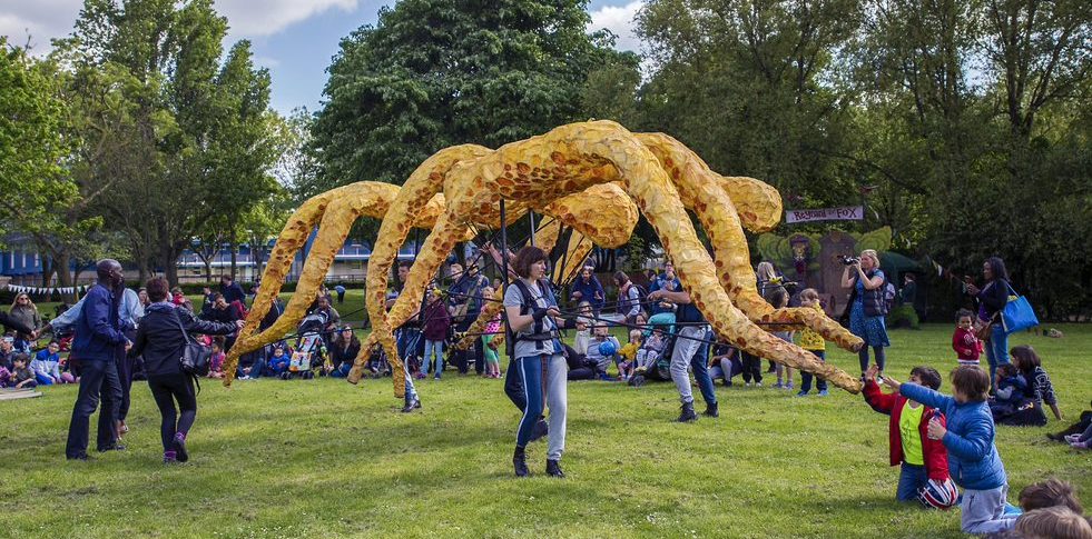 A puppeteer in a park, in front of a crowd, with four squid tentacle puppets. Behind them is another puppeteer, also with four.