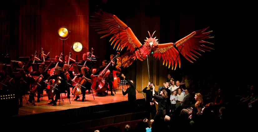 A puppet of a large red bird in between an orchestra and audience, lit by stage lights.