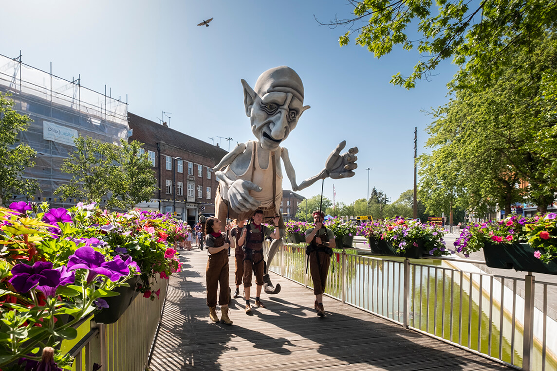Four puppeteers support 'Gnomus' a giant sized puppet of a giant, with grey skin, pointed ears and a large nose. They're on a bridge over the water with flower boxes covering the railings.