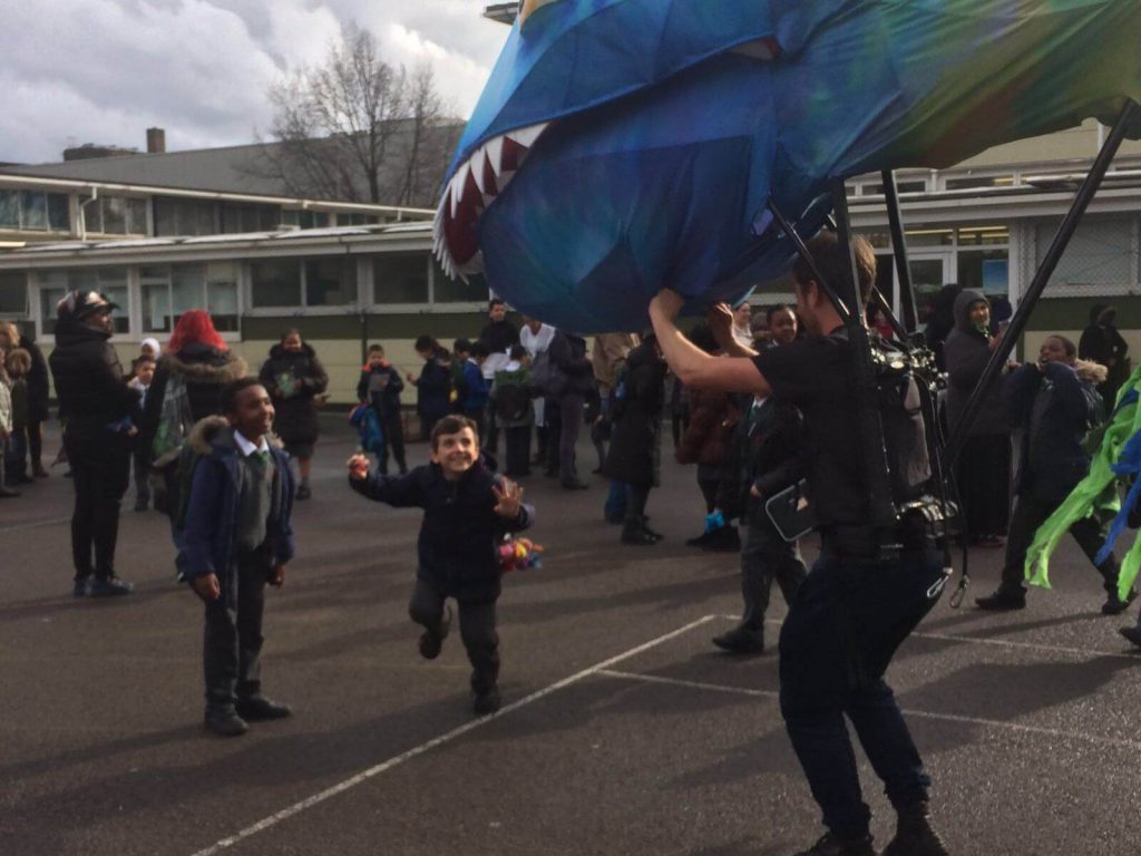 The head of a giant blue and yellow puppet of a monstrous tadpole, supported by a puppeteer. They're in a schoolyard, surrounded by children.