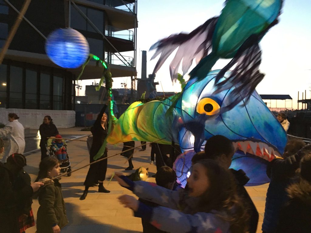 A giant blue puppet of a monstrous tadpole, lit up from the inside. Around it are several children holding smaller lights hanging from rods.