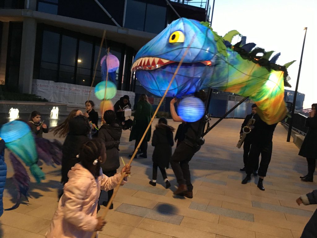 A giant blue puppet of a monstrous tadpole, lit up from the inside. Around it are several children holding smaller lights hanging from rods.