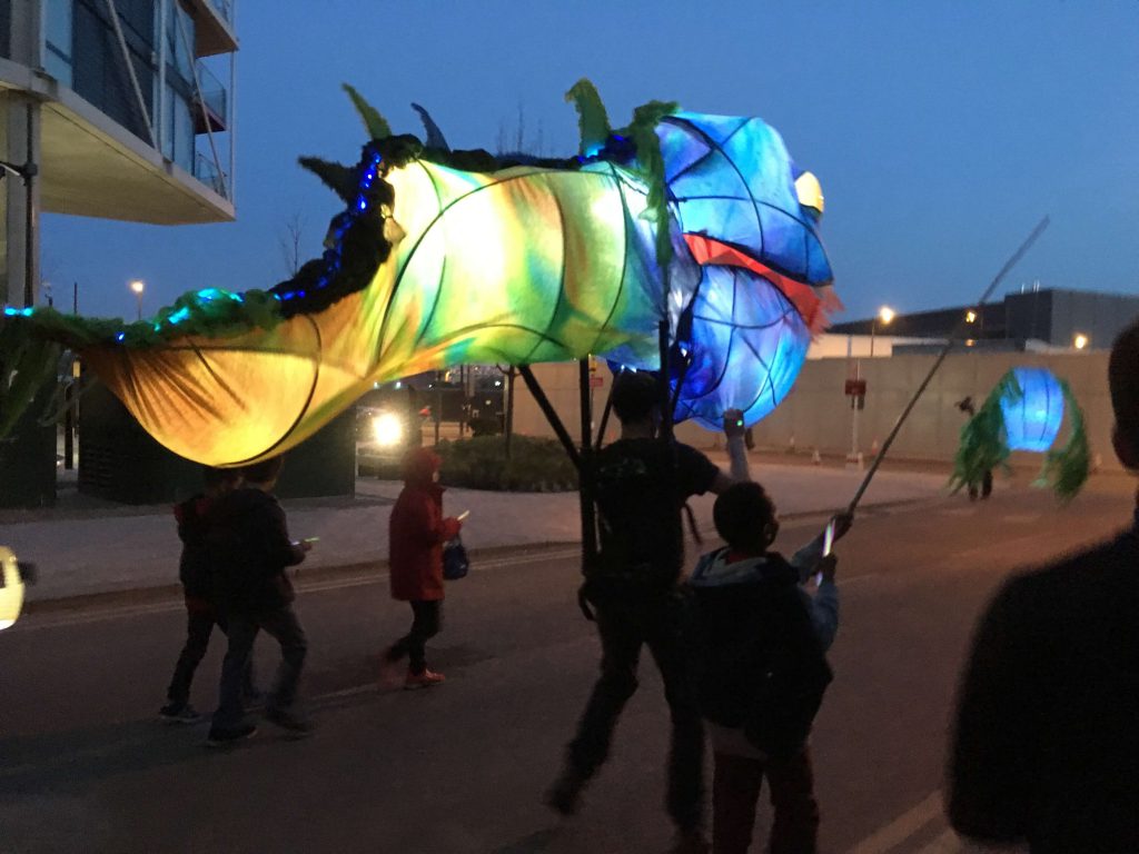 A giant blue and yellow puppet of a monstrous tadpole, lit up from the inside, at night. Around it are several children.