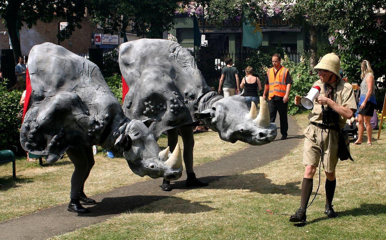 Two large puppets made to look like the front half of two grey rhinos, being worn by puppeteers so that only the puppeteers' legs are visible. The rhinos 'face' a performer dressed like a zookeeper..