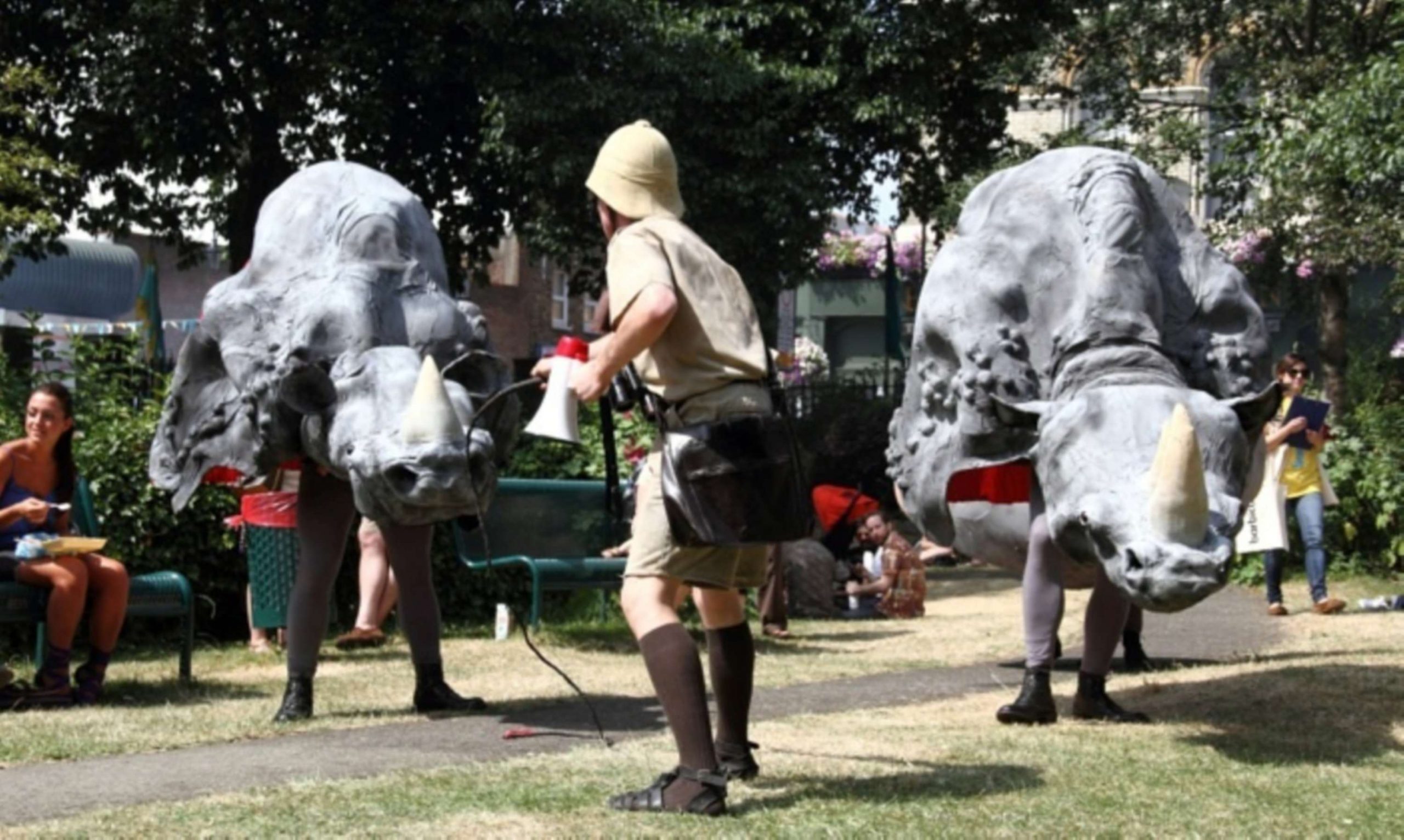 A performer dressed as a zookeeper, facing the puppets - the life-size puppets of the fronts of two rhinos face the camera.