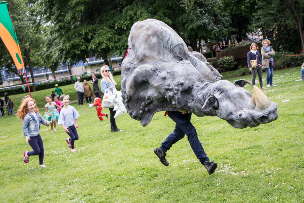 A large puppet made to look like the front half of a rhino. Its legs are the legs of the puppeteer, and they're running away from some laughing children in a park.