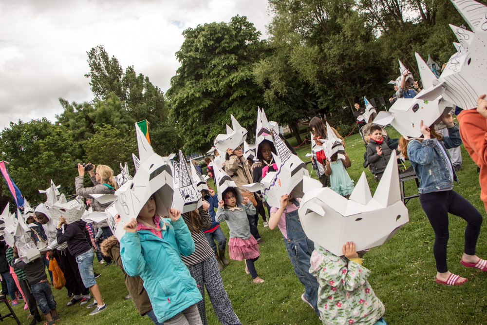 A big group of children in a park, all wearing 3D card rhino masks that they have decorated.