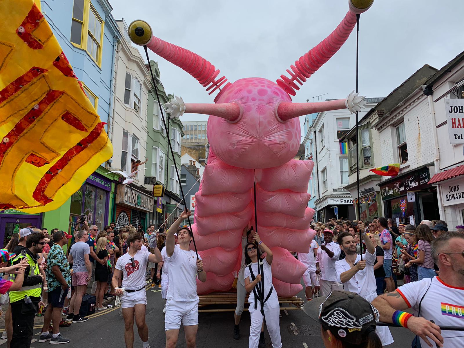 Numerous puppeteers support a giant puppet of a pink snail with a rainbow shell, moving it through a crowded street.