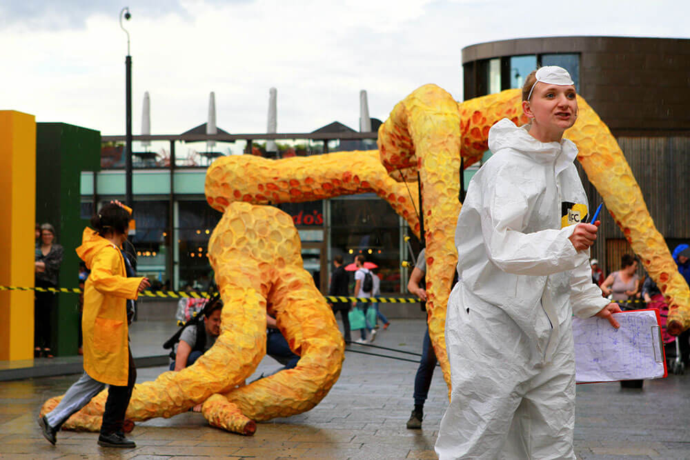 Two people, one in a yellow-orange coat, one in white overalls with a medical mask on their forehead and holding a clipboard. They're both in front of several giant orange puppets of squid tentacles.