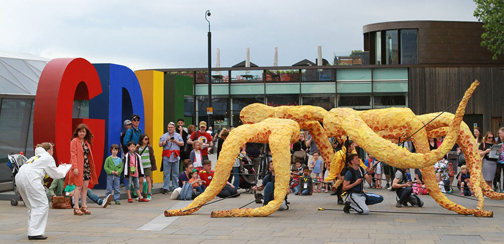 Eight models of orange squid legs, supported by puppeteers, in front of a crowd and a building. A person in white overalls stands in front of them, holding a clipboard.