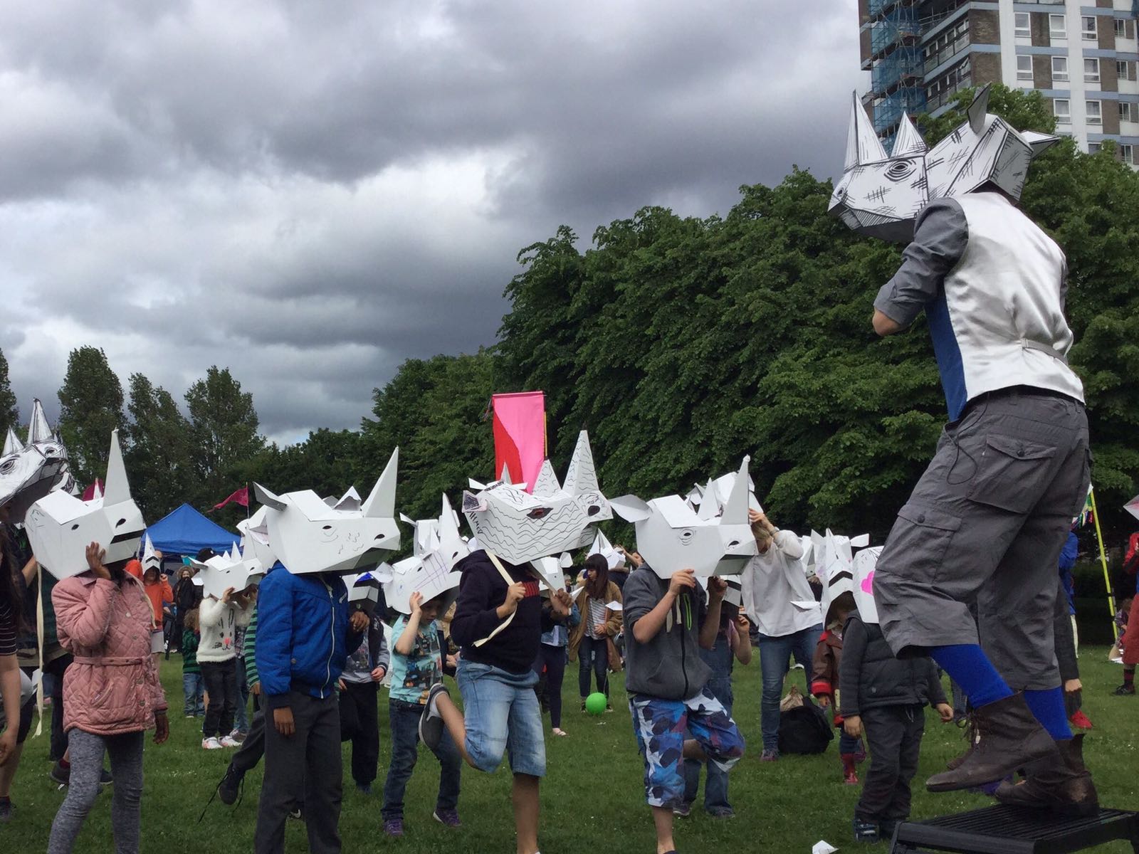 A performer in a rhino mask in front of a crowd of children wearing white 3D rhino masks which they have decorated. They're in a park.