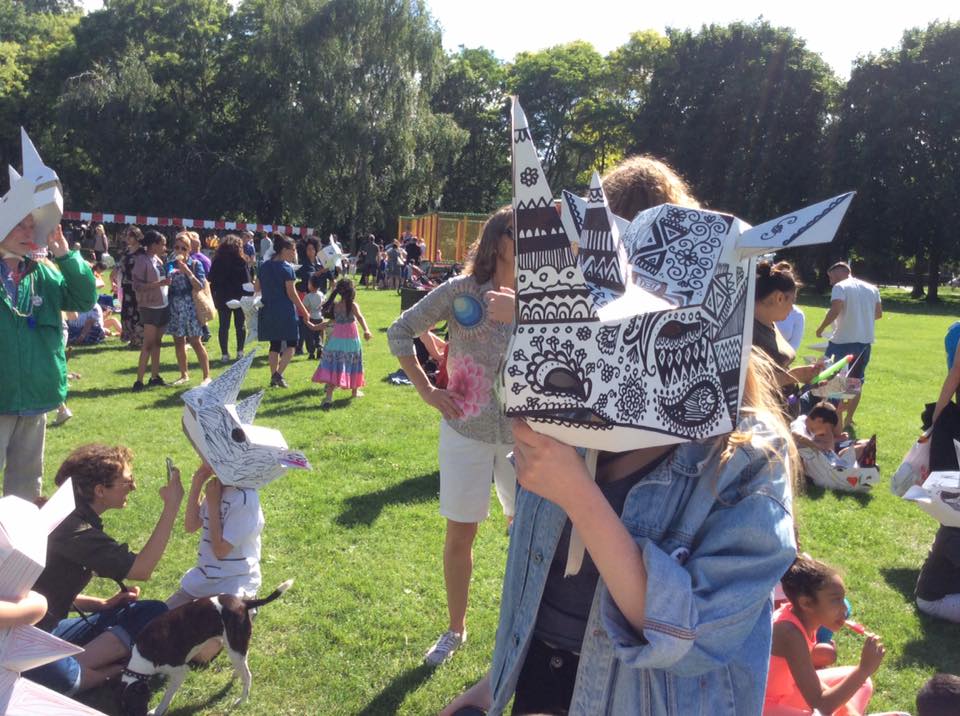 A crowd in a park, several wearing rhino masks. The focus is on someone wearing a 3D rhino mask decorated with intricate patterns.
