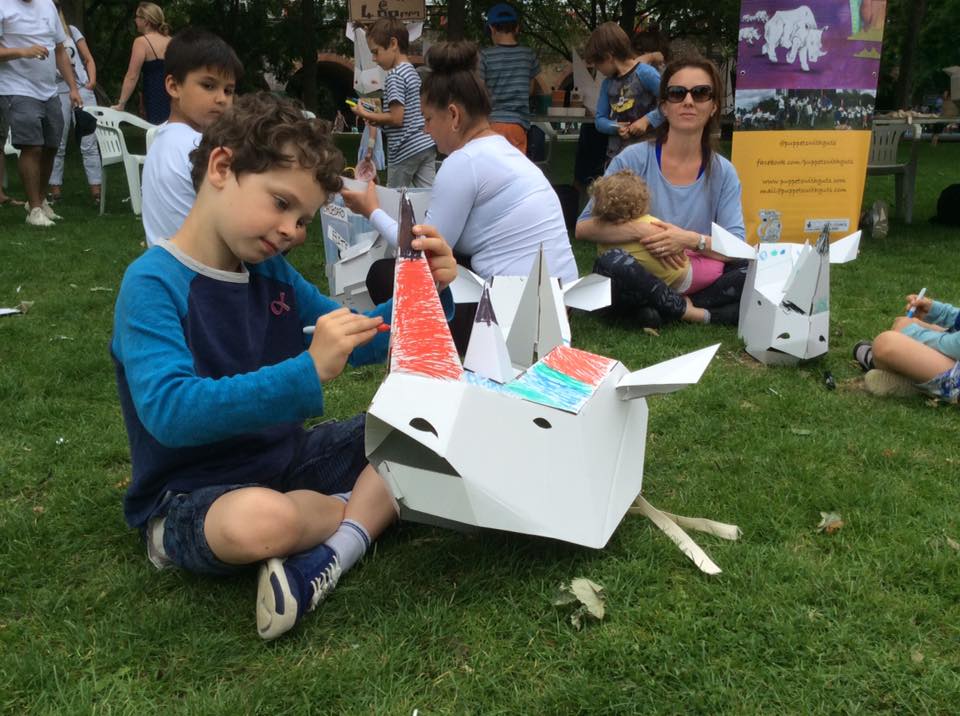 A child sitting on the grass, decorating a 3D card rhino mask with a red pen.