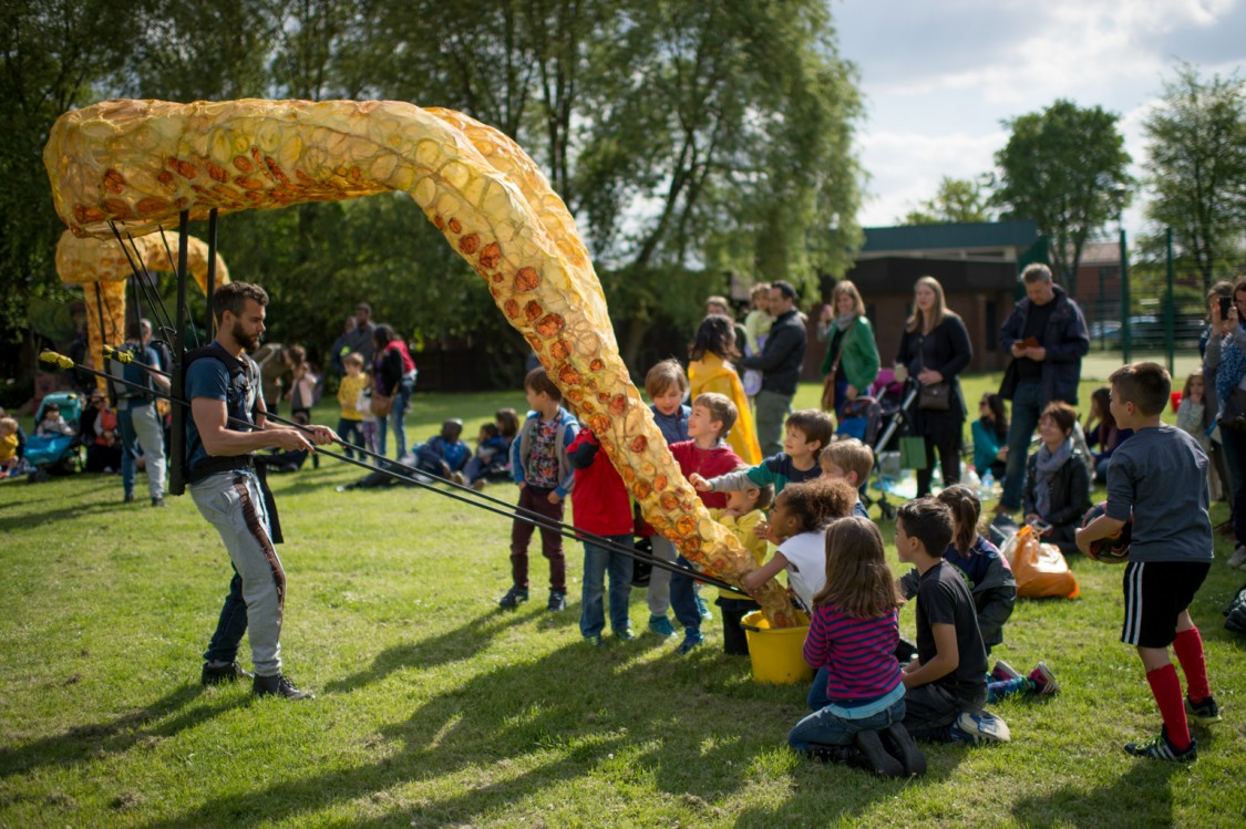 A crowd of children on the grass, reaching out to touch two yellow-orange puppets of giant squid tentacles, both controlled by one puppeteer.