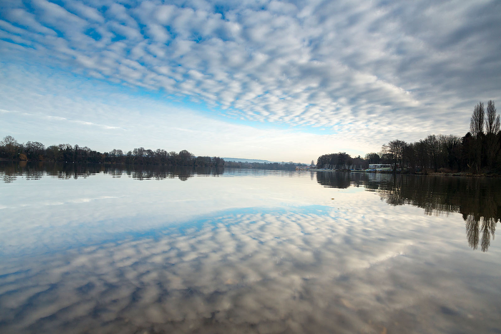 A lake takes up the lower half of the frame and reflects the other half of the image: the cloudy sky, and far away small forests.