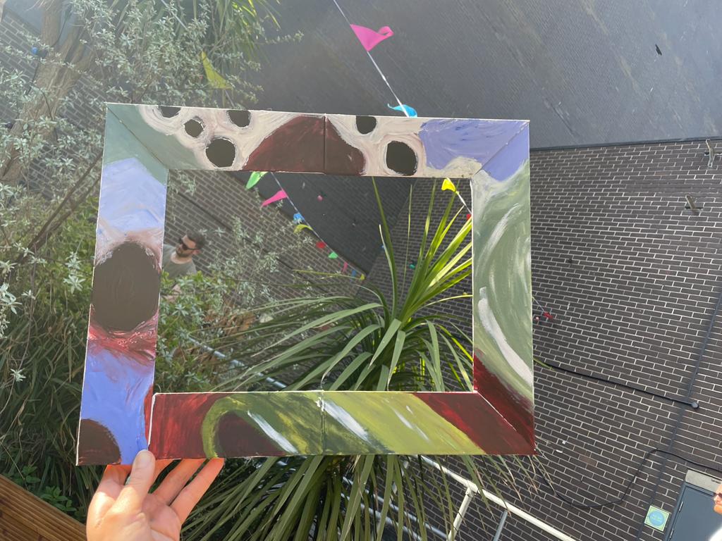 A cardboard picture frame, painted with abstract patterns in blue, brown, green, black and white. It's held up so that it frames some plants, bunting, a wall, and a man wearing sunglasses.