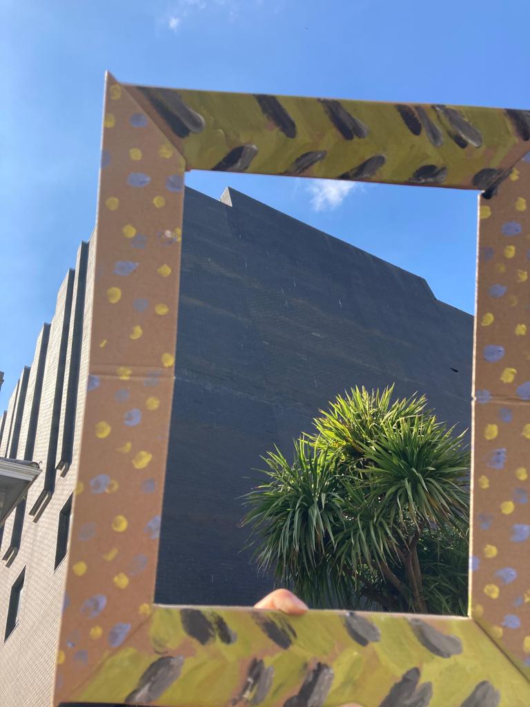 A cardboard picture frame, painted with yellow and blue dots at the sides and green/black patterns at the top and bottom, held up so that it is framing the back of a building, the sky, and a palm tree.