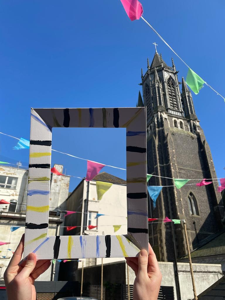 A white cardboard picture frame, painted with thin stripes of yellow, blue, black, and green paint. Hands hold it up in front of an old church spire and many white houses, with bunting strung up - the frame frames the back of a house with some bunting. The sky is clear and bright blue.